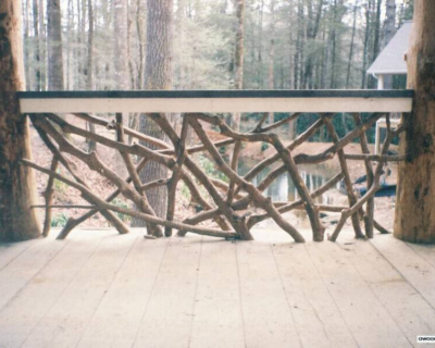 Deck with dense woven twig railing and log posts near forest.