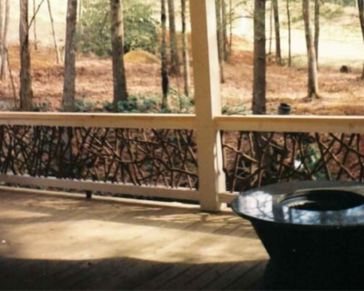 Wooden porch with handcrafted woven branch railing overlooking a wooded forest area, with a black fire pit in the foreground.