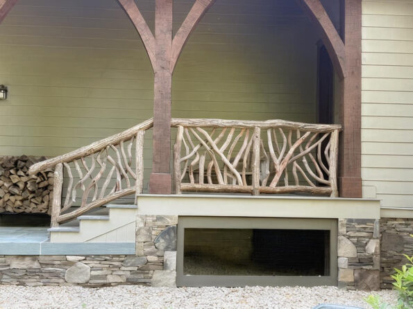 Rustic porch railing made of intertwined branches with a stone foundation and stacked firewood nearby.