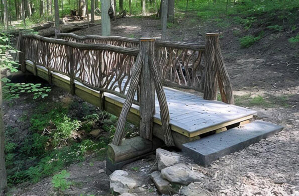 Wooden footbridge with decorative branch-style railings crossing forest ravine surrounded by trees.