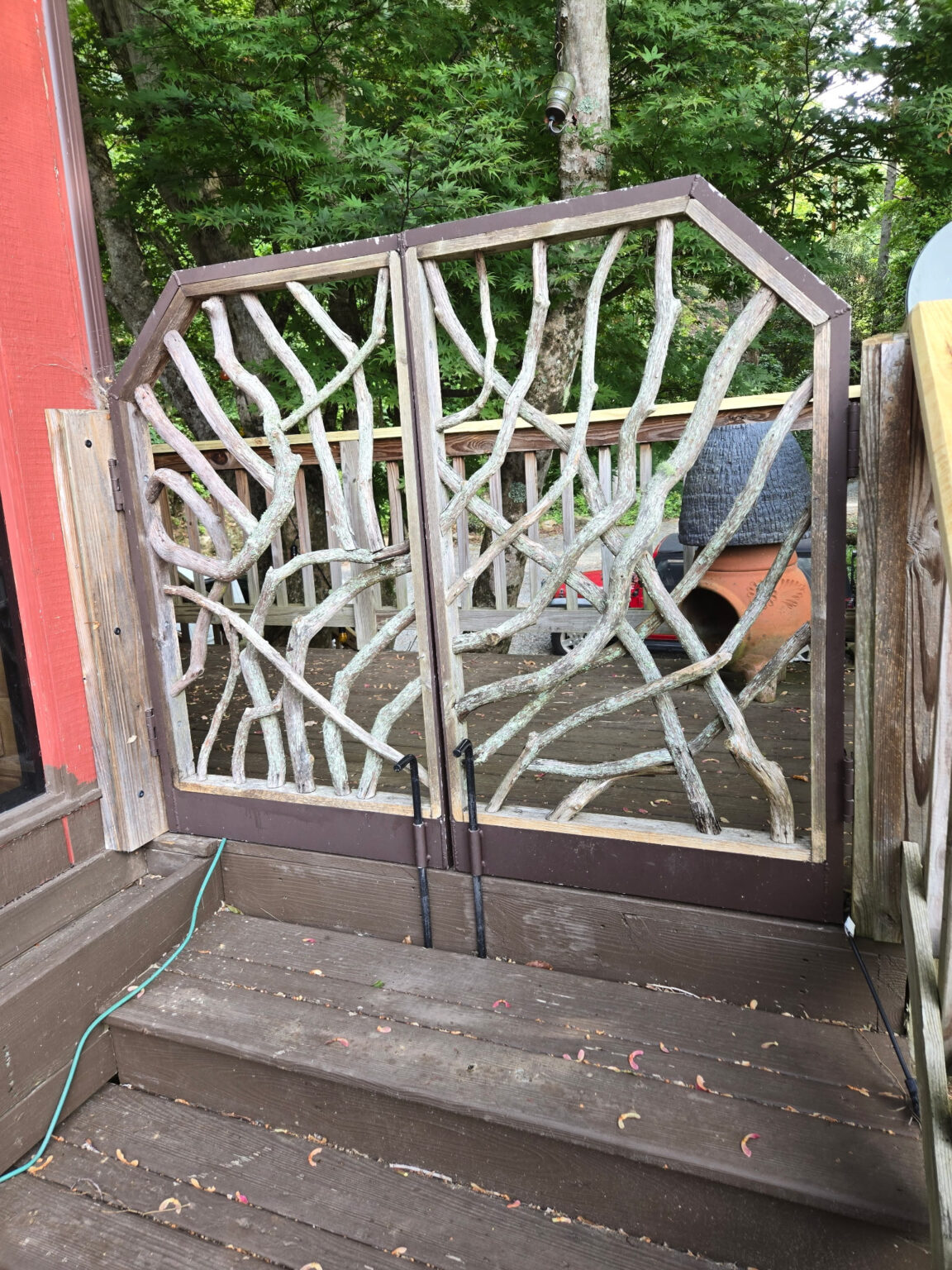 Rustic wooden porch gate made from mountain laurel branches on a deck surrounded by trees and outdoor furniture.
