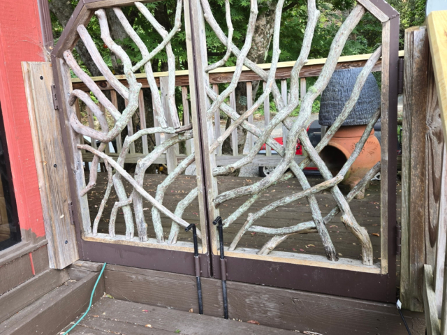 Rustic wooden porch gate made from mountain laurel branches on a deck surrounded by trees and outdoor furniture.