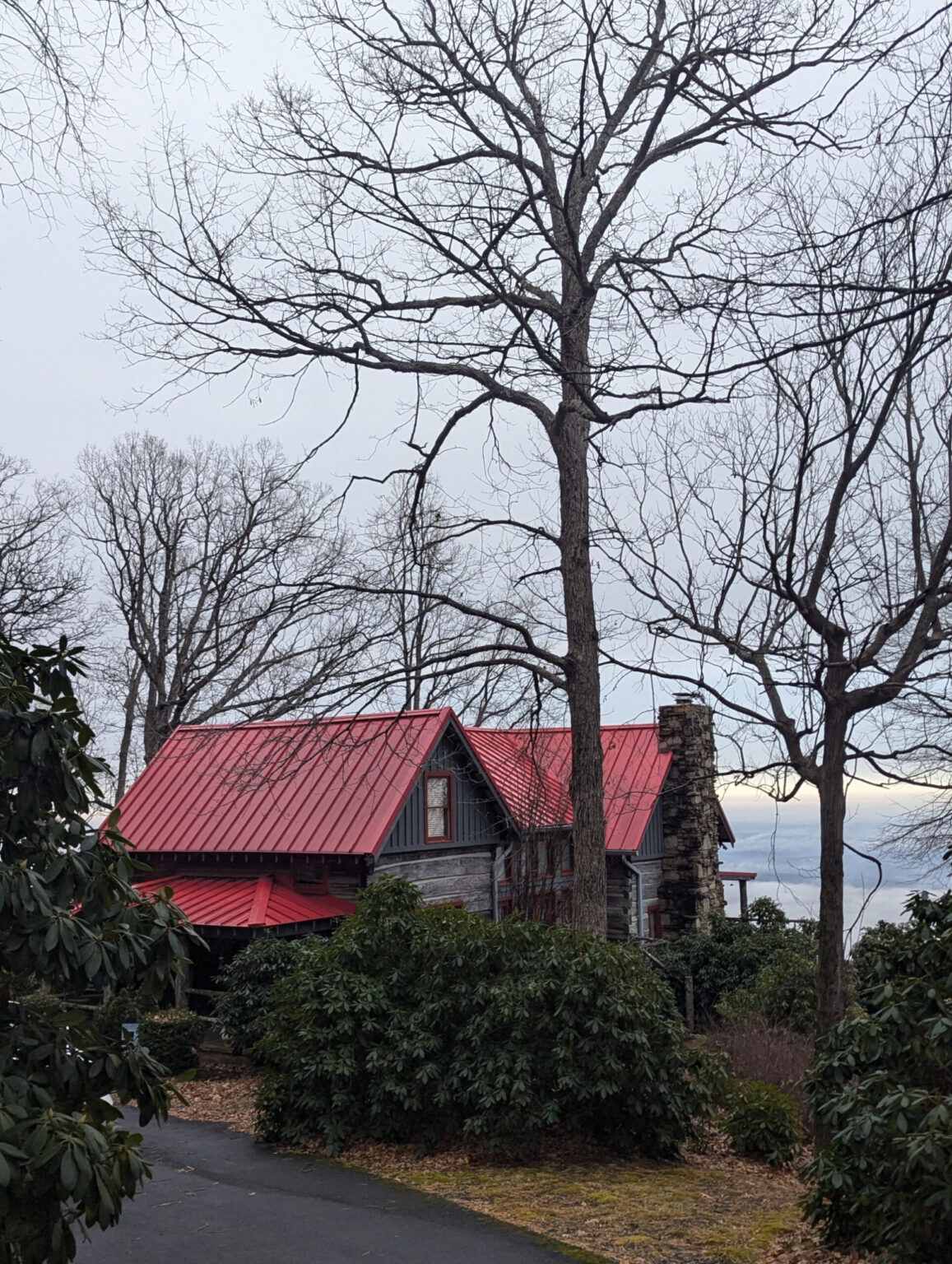 Log cabin with red metal roof and stone chimney surrounded by bare trees and green bushes.