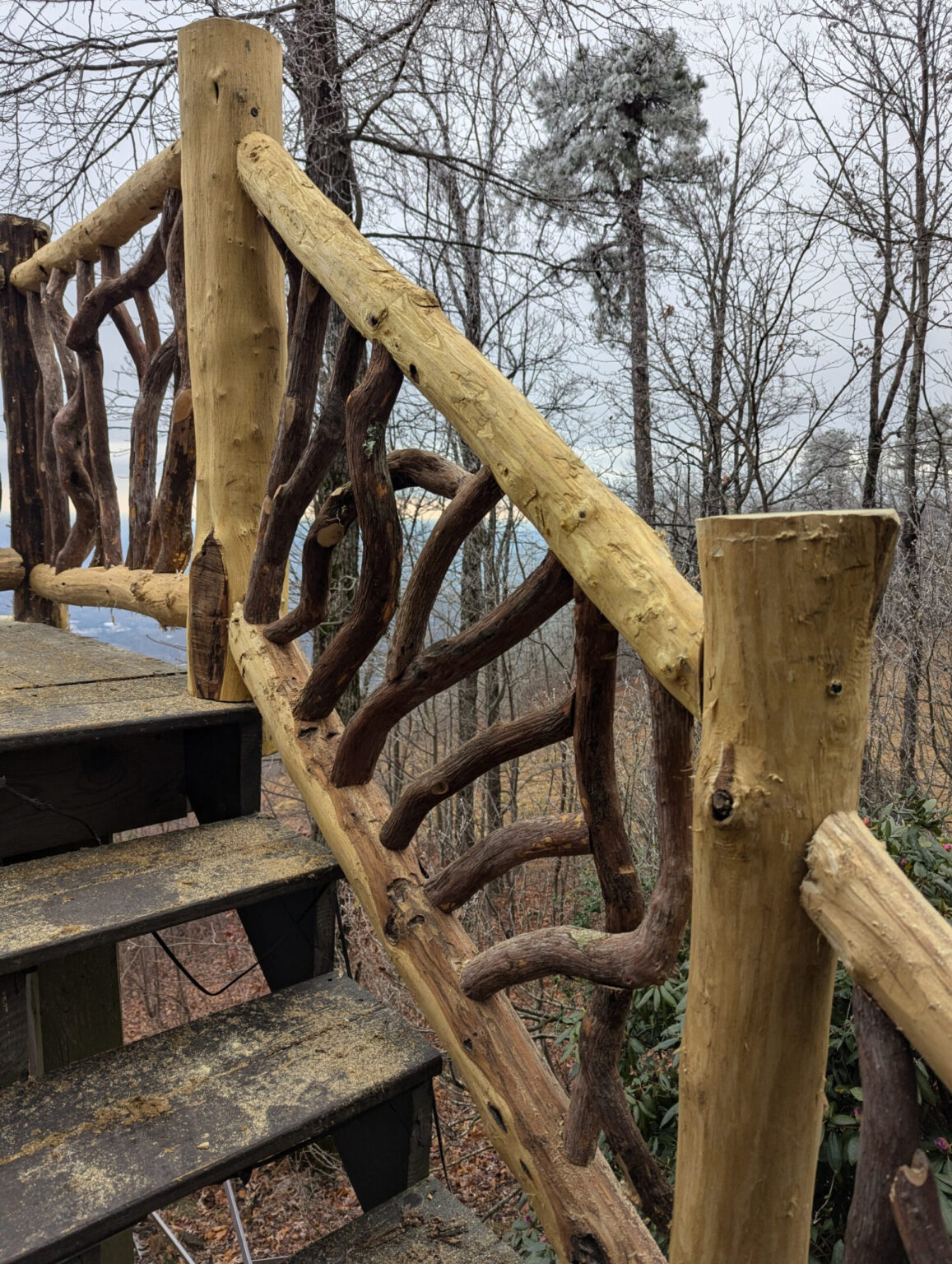 Rustic wooden staircase with intricate woven branch railing overlooking a forest.