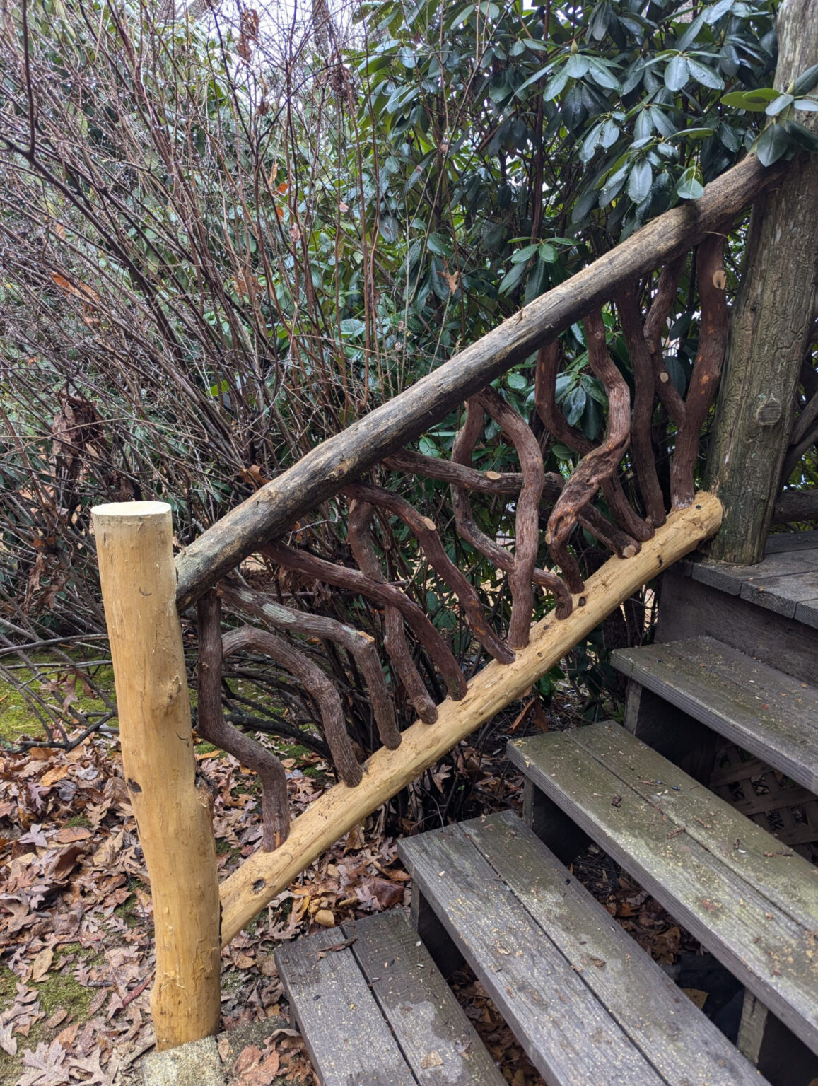 Rustic wooden staircase with woven branch railing surrounded by dense bushes and fallen leaves.