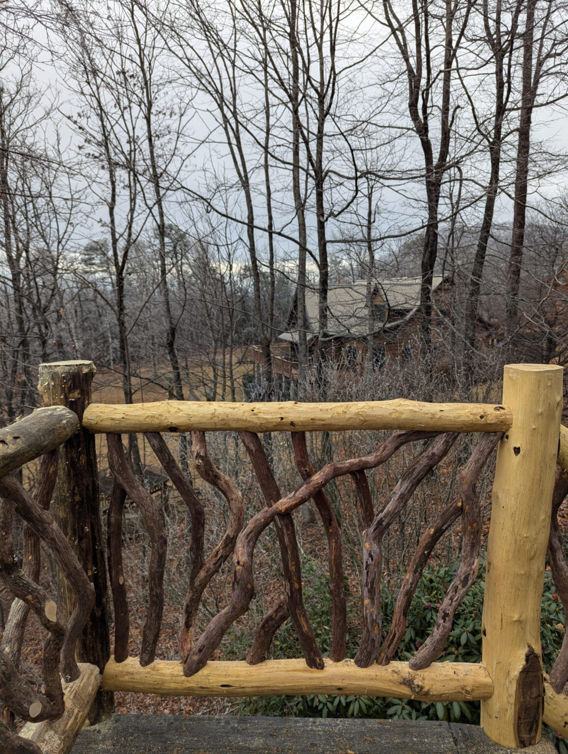 Rustic woven branch railing on a wooden deck overlooking a forest of bare trees and a wooden cabin in the distance under an overcast sky.