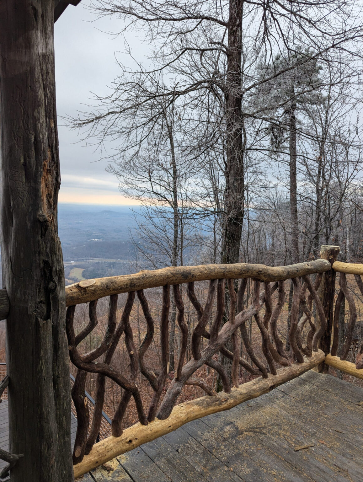 Rustic woven branch railing on a wooden deck overlooking a vast valley and distant mountains through bare winter trees under an overcast sky.