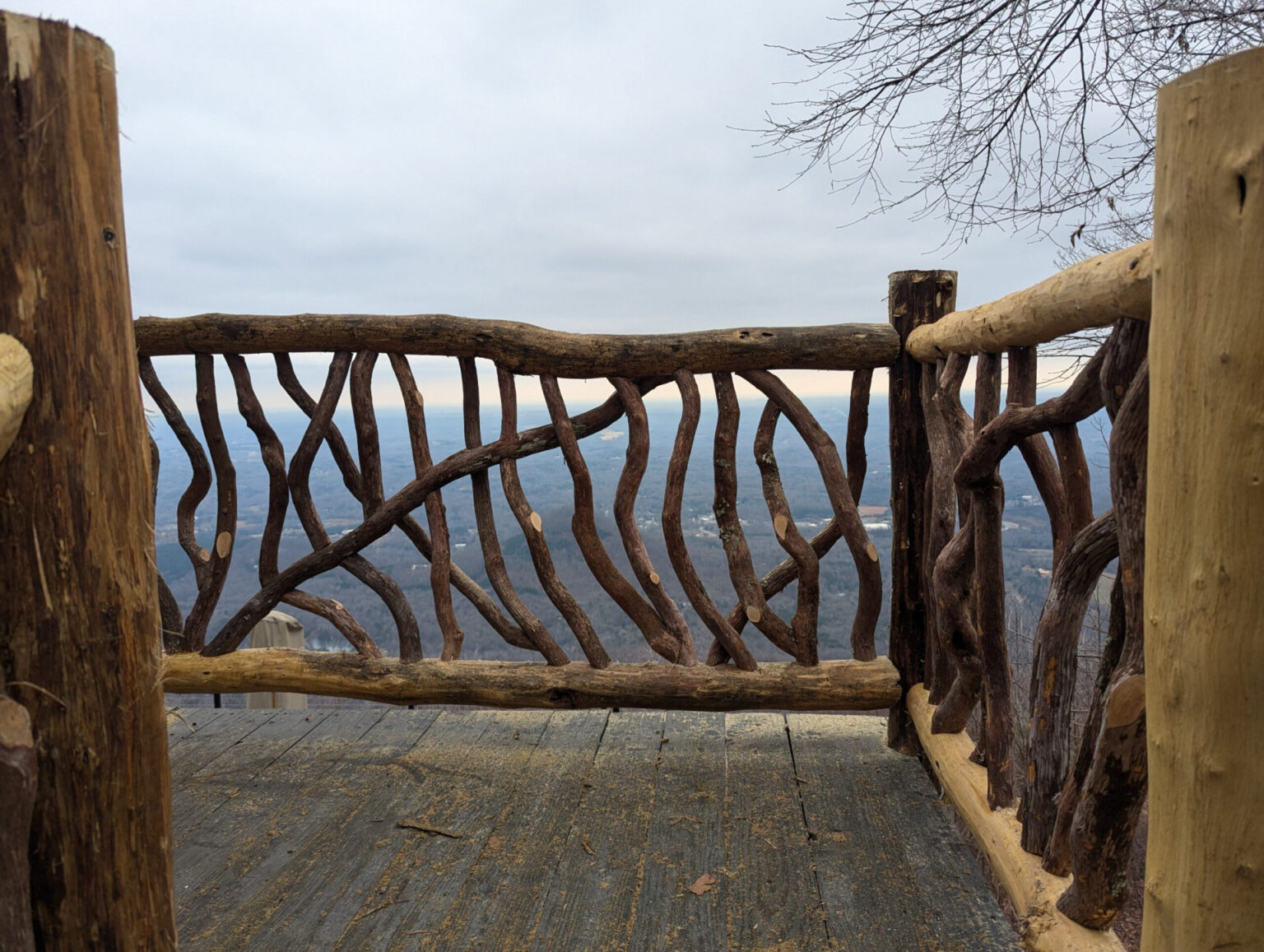 Rustic woven branch railing on a wooden deck overlooking a vast winter landscape with hazy hills under an overcast sky.