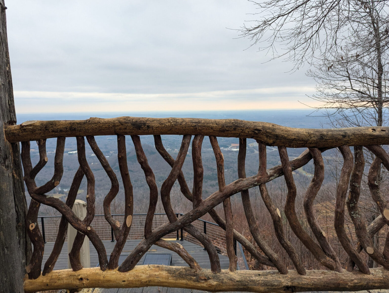 Rustic woven branch railing on a wooden deck overlooking a vast hazy valley and distant hills under an overcast winter sky.