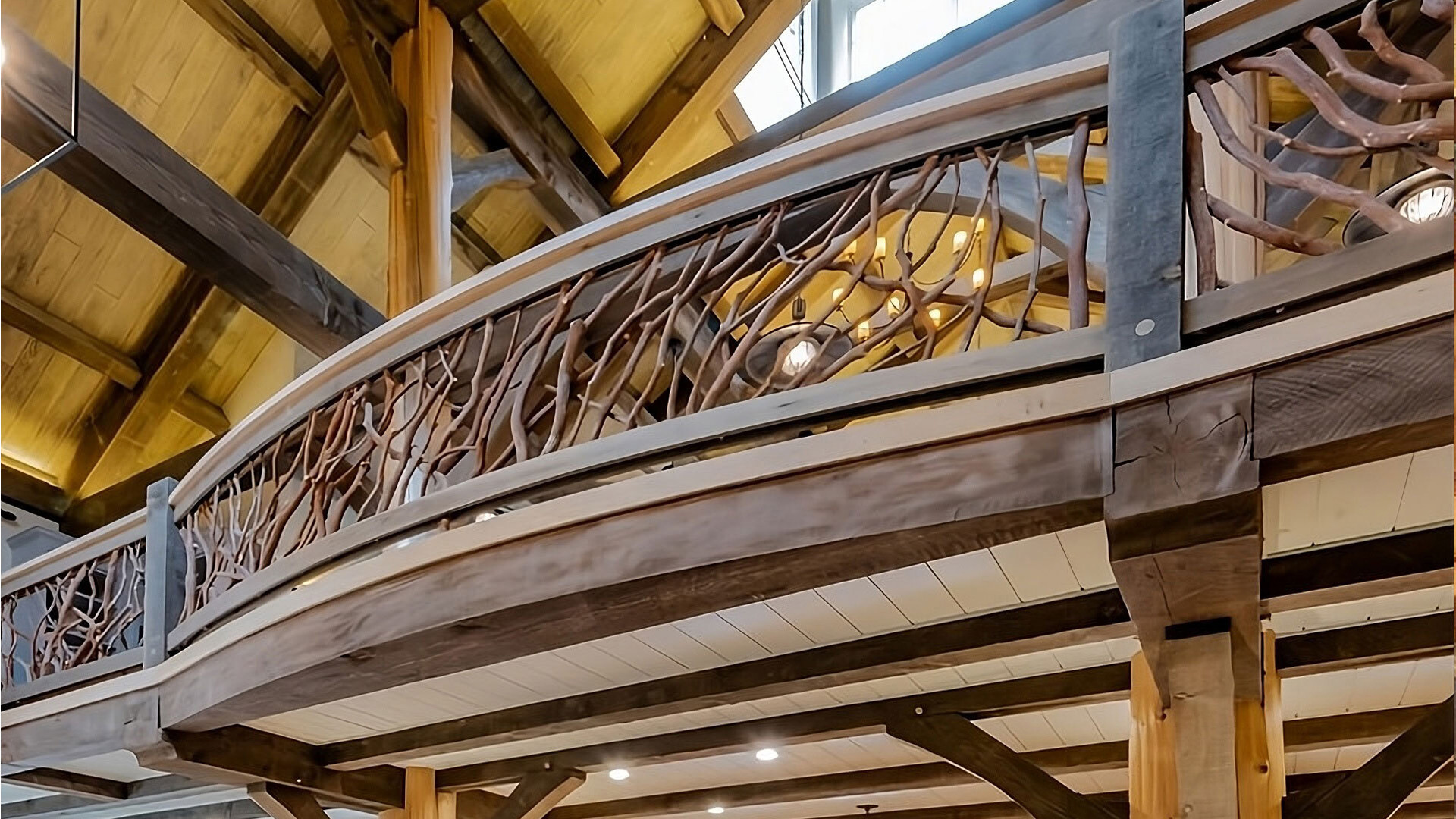 Curved wooden loft balcony with intricate handcrafted woven branch railing inside a spacious log home with high vaulted ceilings and exposed beams.