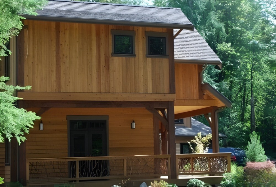 Two-story wooden house with vertical siding, a covered deck featuring handcrafted woven branch railing, surrounded by lush green trees.
