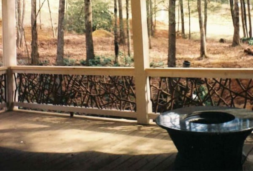 Wooden porch with handcrafted woven branch railing overlooking a wooded forest area, with a black fire pit in the foreground.
