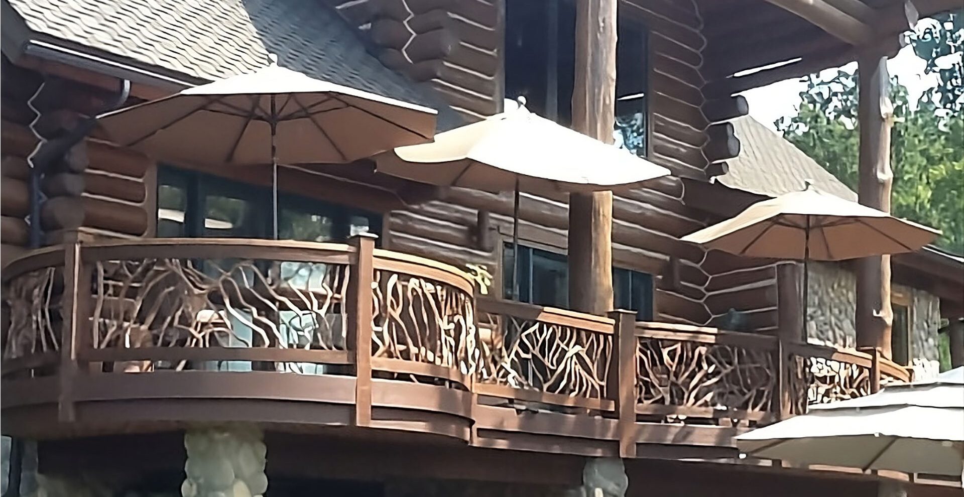 A rustic log cabin lodge with a wooden balcony featuring branch-style railings and several beige patio umbrellas, surrounded by trees in a natural outdoor setting.