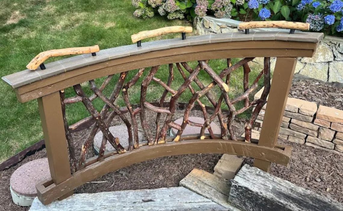 A decorative wooden garden bridge with twig railings and curved handrails, set in a landscaped yard.