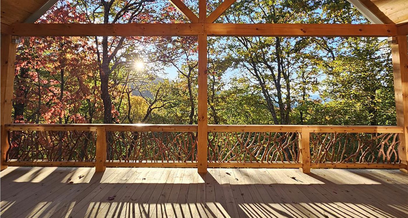 Covered wooden deck with handcrafted woven branch railing overlooking a vibrant autumn forest with colorful red, orange, and green trees under a bright sun.