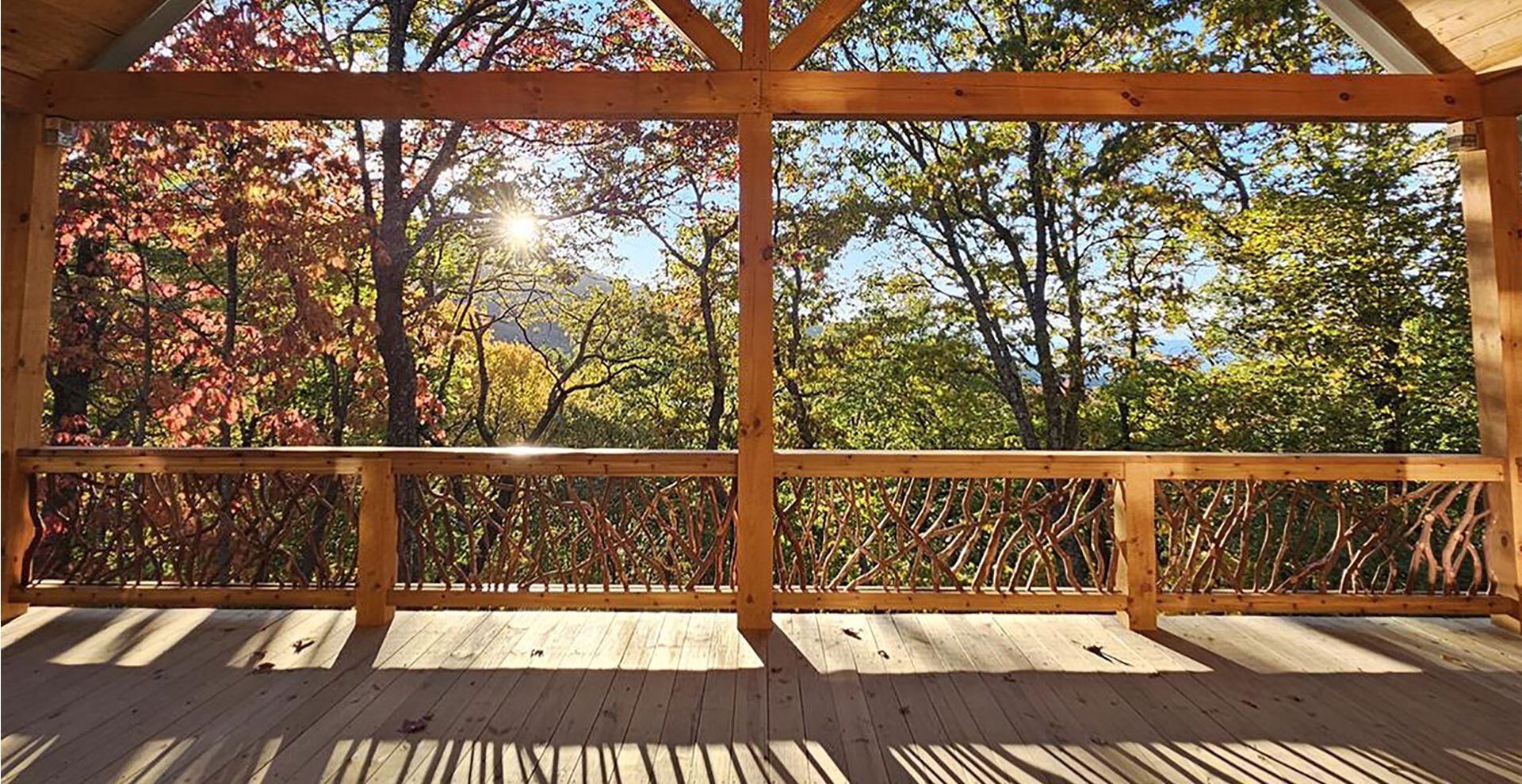 Covered wooden deck with handcrafted woven branch railing overlooking a vibrant autumn forest with colorful red, orange, and green trees under a bright sun.