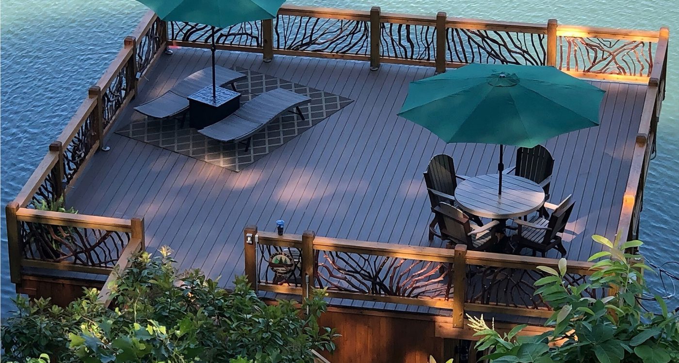 Spacious lakeside wooden deck with handcrafted woven branch railing, lounge chairs, dining set, and green umbrellas overlooking the water.