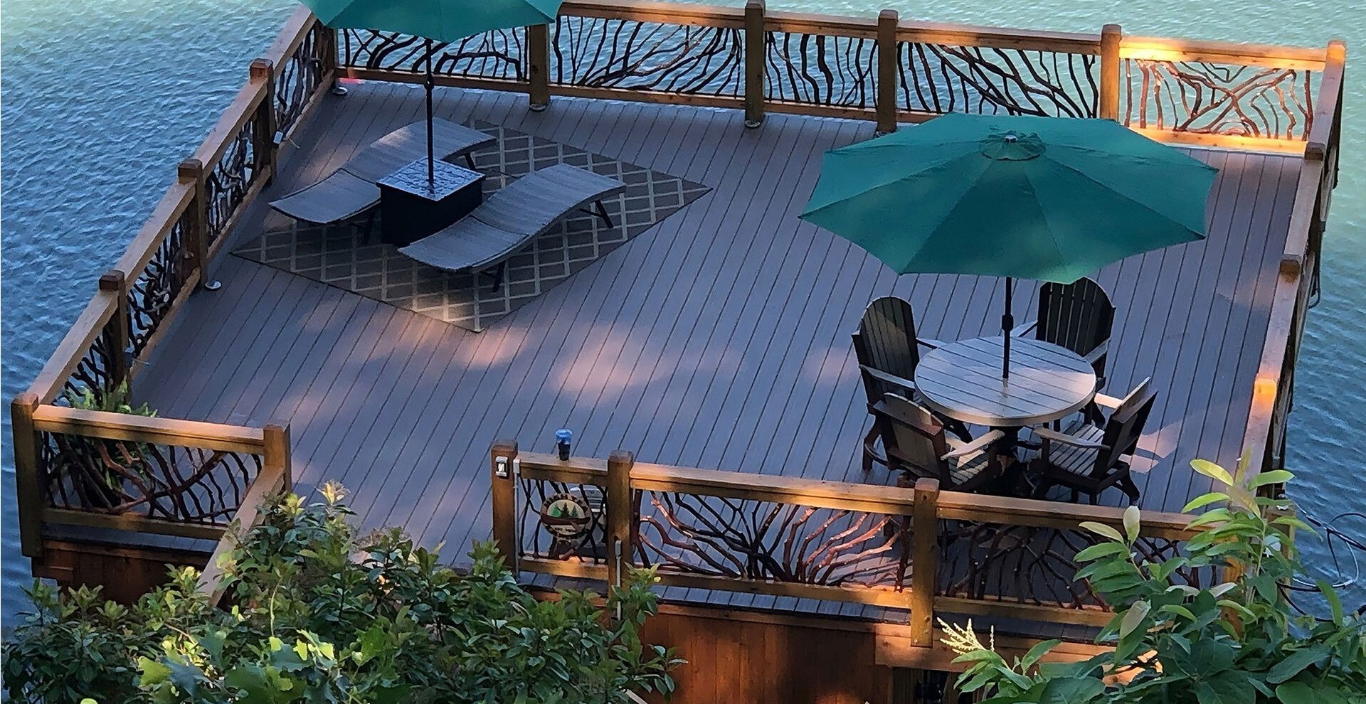 Spacious lakeside wooden deck with handcrafted woven branch railing, lounge chairs, dining set, and green umbrellas overlooking the water.