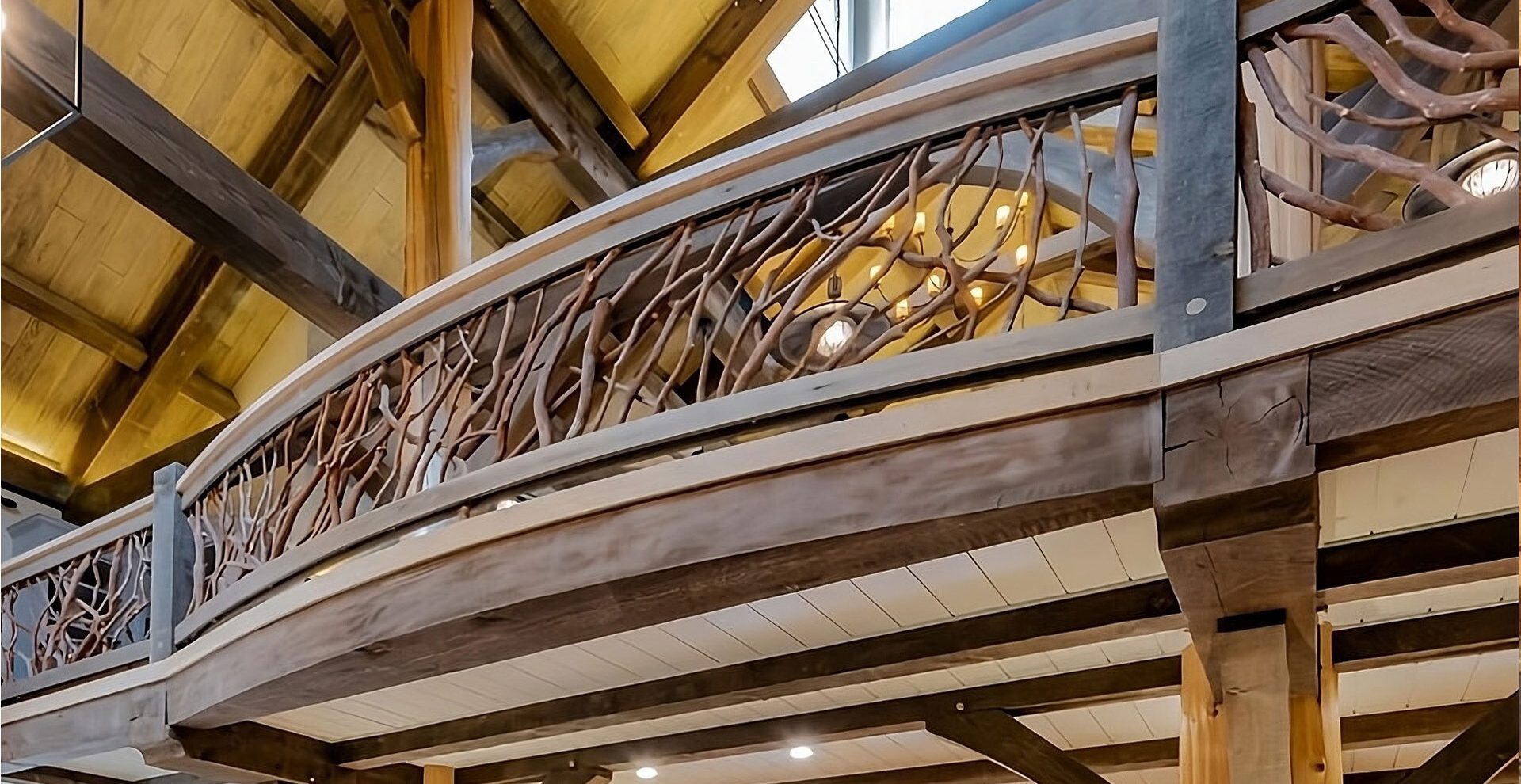 Curved wooden loft balcony with intricate handcrafted woven branch railing inside a spacious log home with high vaulted ceilings and exposed beams.