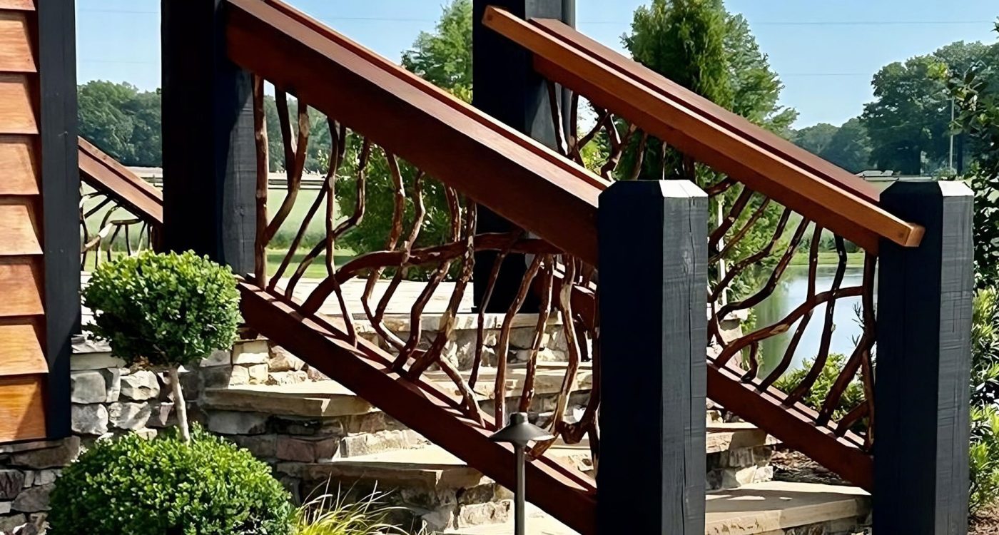 Wooden staircase with handcrafted woven branch railing in rich brown tones, set against stone steps, a lake, and green trees under a clear blue sky.