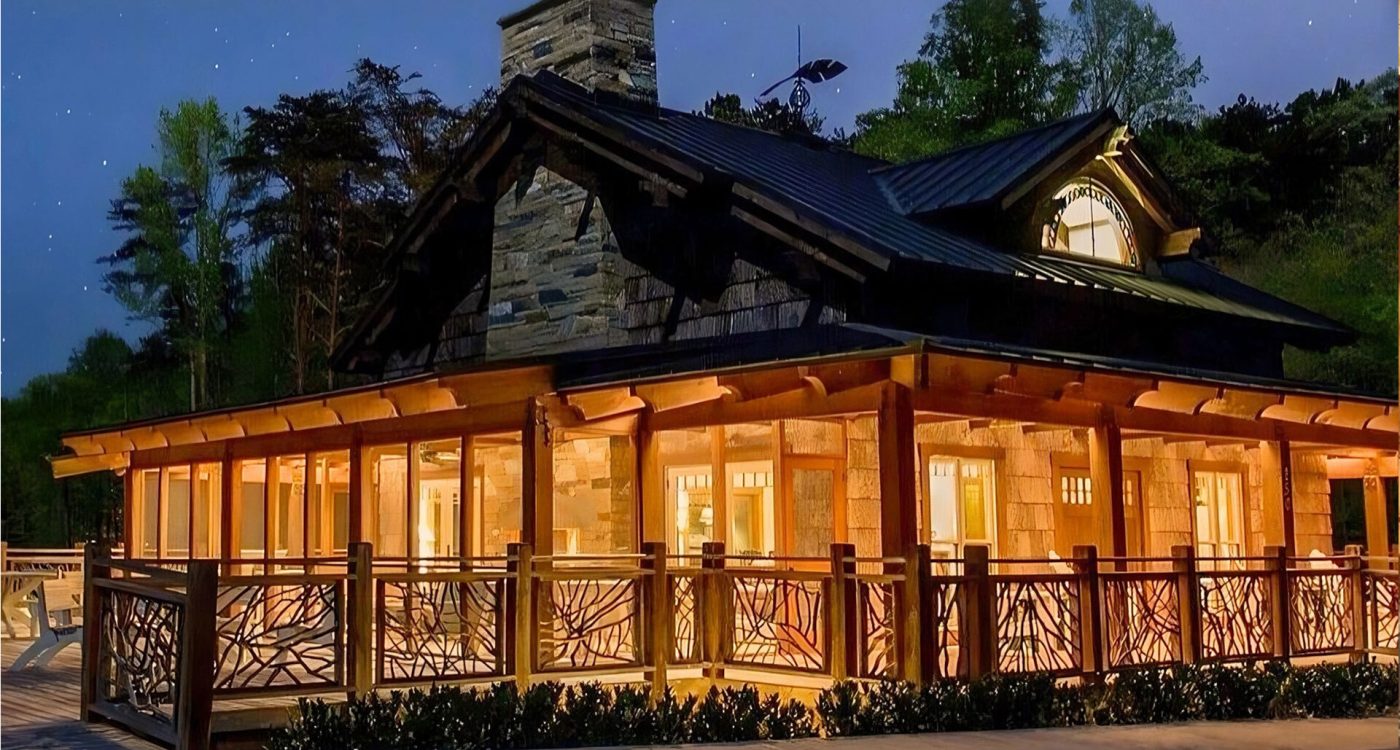 Large log and stone house at night with warm interior lighting, featuring a wraparound deck with handcrafted woven branch railings under a starry sky.