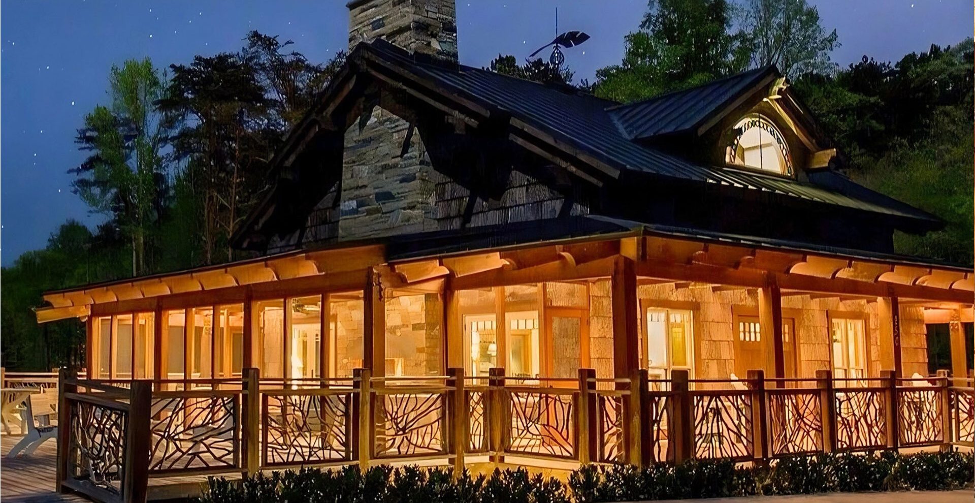 Large log and stone house at night with warm interior lighting, featuring a wraparound deck with handcrafted woven branch railings under a starry sky.