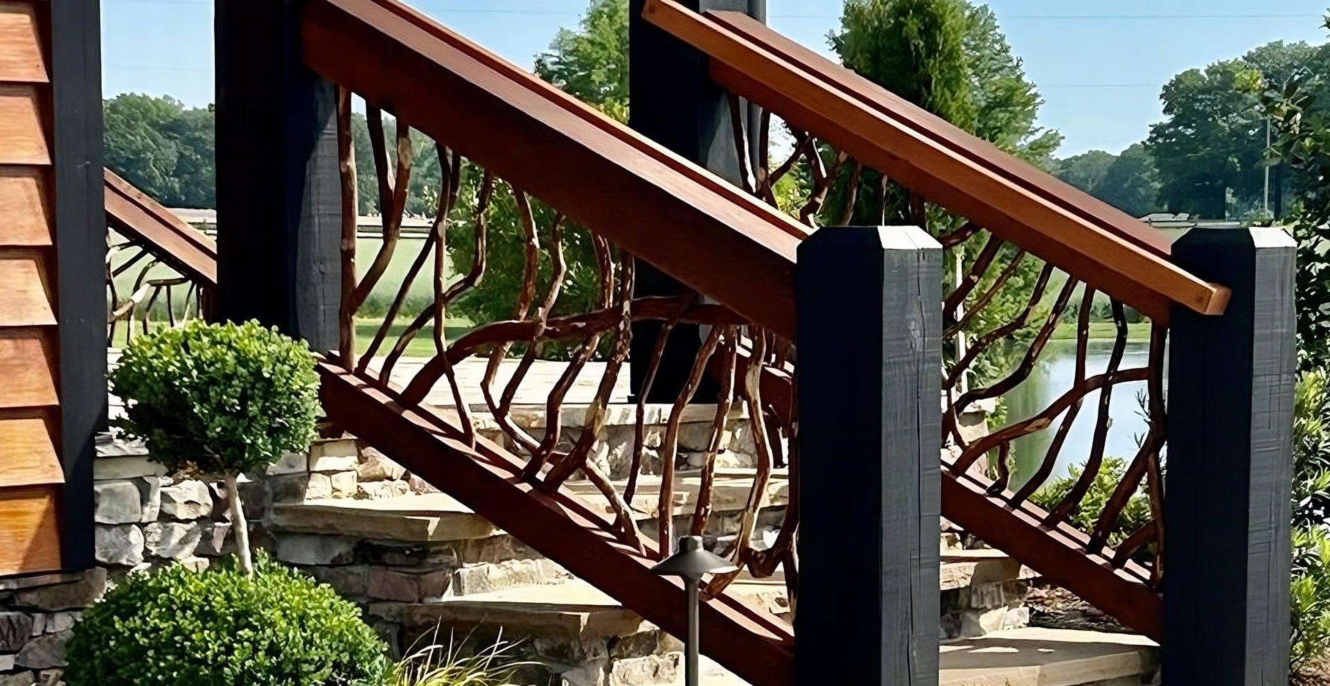 Rustic wooden stair railing with branch-style balusters beside stone steps in a landscaped yard.