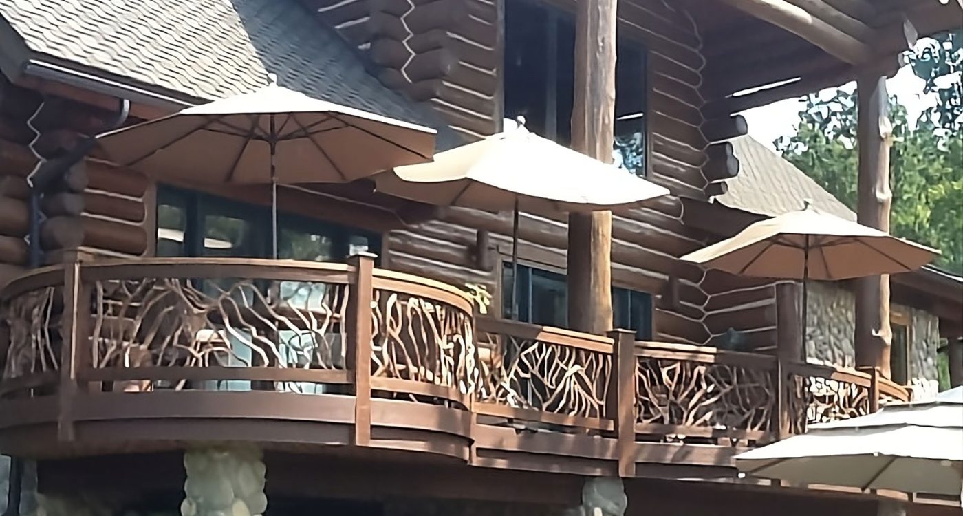 Log cabin home with a curved wooden deck featuring rustic branch-style railings and patio umbrellas.