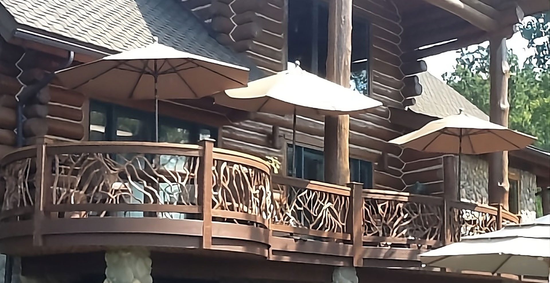 Log cabin home with a curved wooden deck featuring rustic branch-style railings and patio umbrellas.