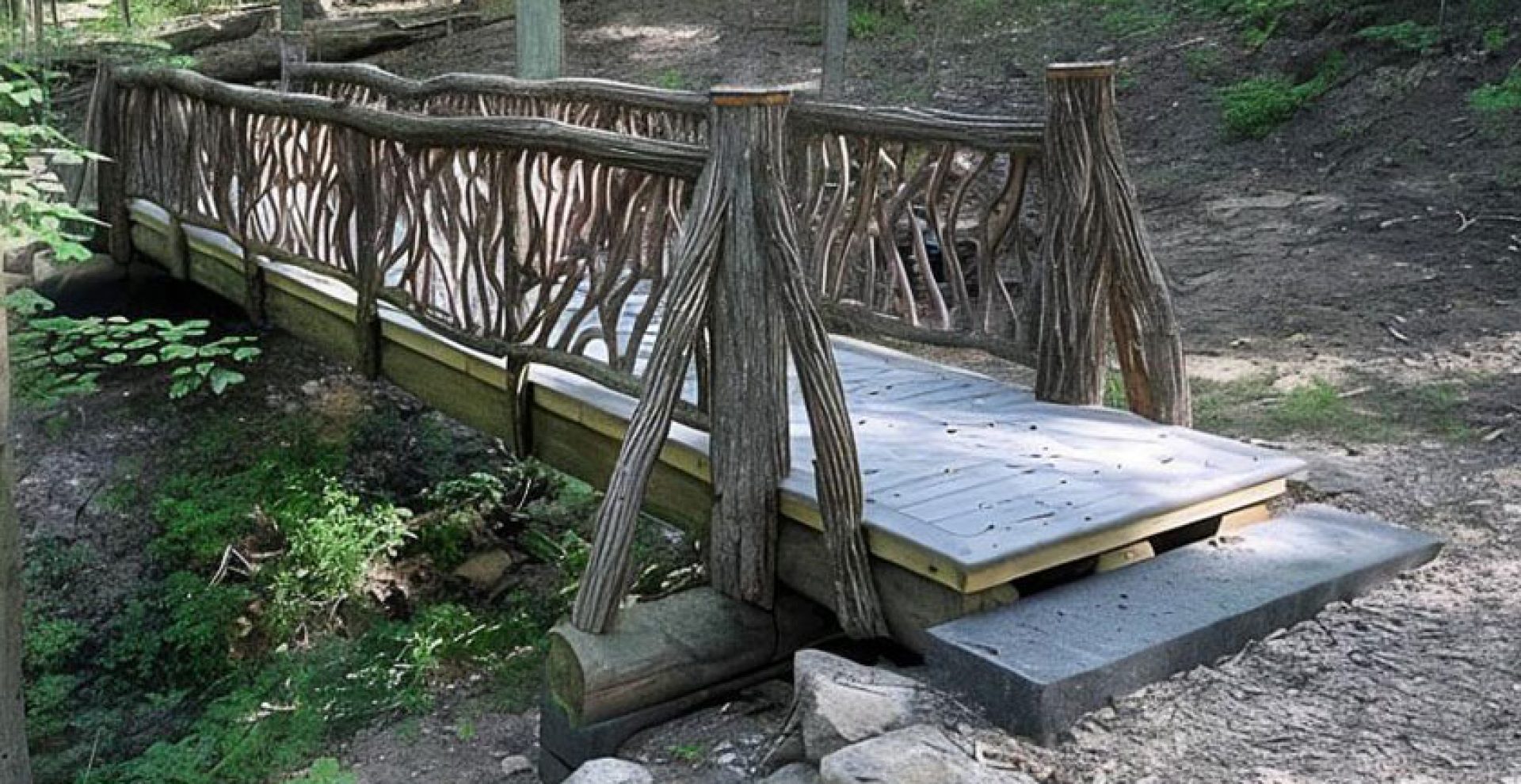 Wooden footbridge with decorative branch-style railings crossing forest ravine surrounded by trees.