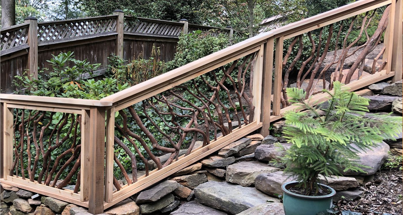 Wooden bridge or walkway with handcrafted woven branch railing, set over a stone retaining wall with lush greenery and a potted evergreen in the foreground.