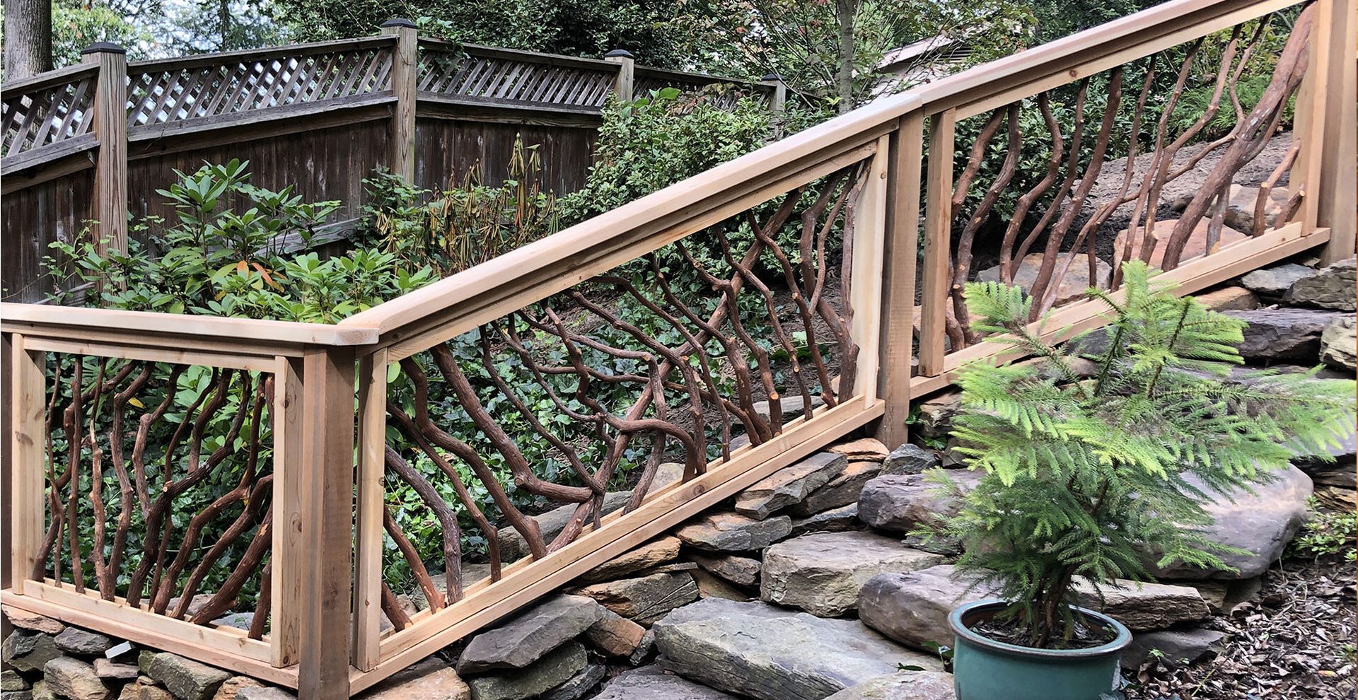 Wooden bridge or walkway with handcrafted woven branch railing, set over a stone retaining wall with lush greenery and a potted evergreen in the foreground.