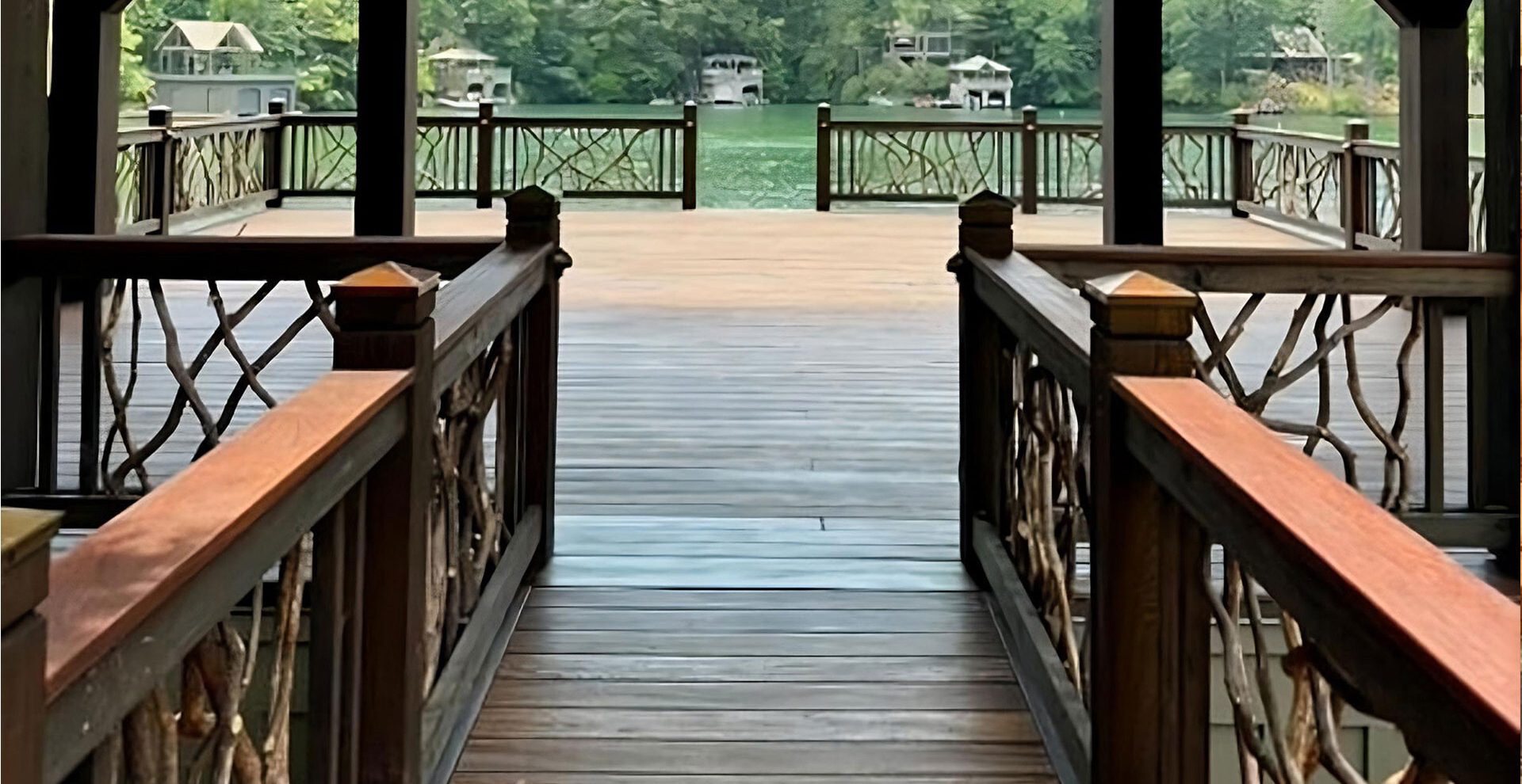 Spacious wooden deck over water with handcrafted woven branch railing, featuring lounge chairs, a dining set with green umbrellas, and views of the lake.