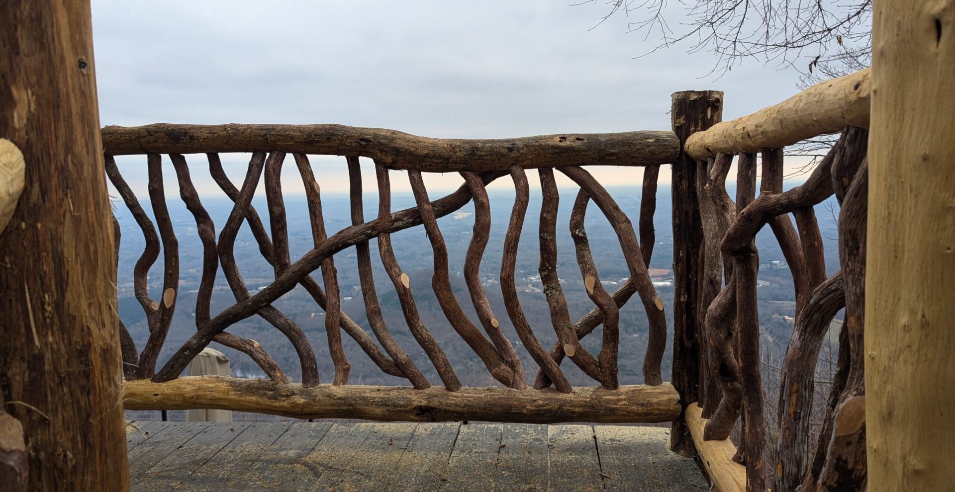 Rustic woven branch railing on a wooden deck overlooking a vast winter landscape with hazy hills under an overcast sky.