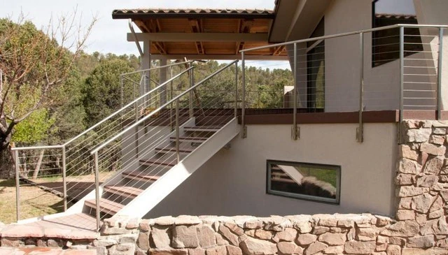 A minimal contemporary cable railing design with silver cables strung between small diameter square metal pipes and a round pipe, enhancing the aesthetic of deck stairs.