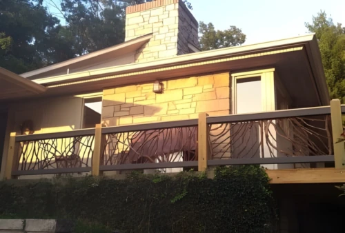 A house with stone and wood exterior featuring a second-floor deck with beautiful handcrafted woven branch railings, surrounded by trees and greenery.