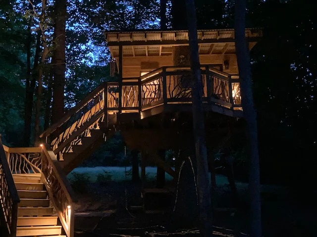 Front view of a custom treehouse featuring mountain laurel handrails, elegantly illuminated at nighttime, creating a captivating outdoor scene.