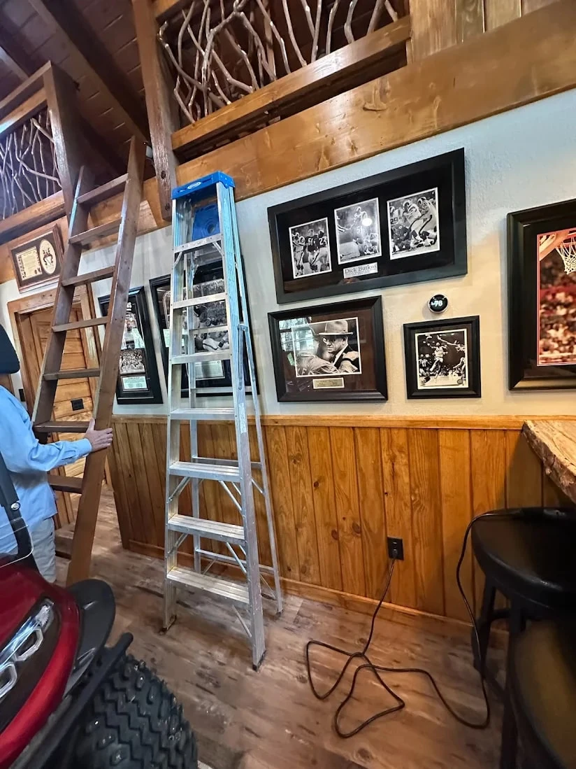 Wooden ladder and metal step ladder beneath a loft with branch railing and framed photos on a wood-paneled wall.