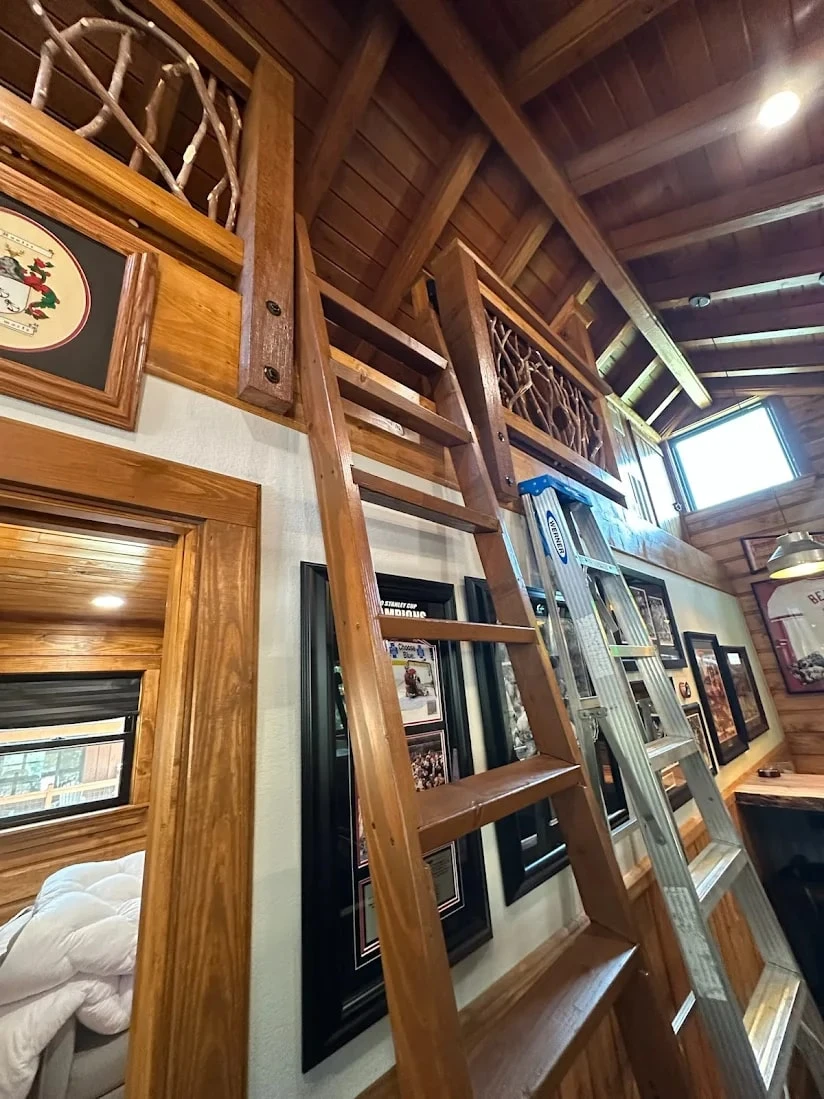 Wooden ladder leading to a loft with branch-style railing in a rustic cabin interior with exposed wood beams.