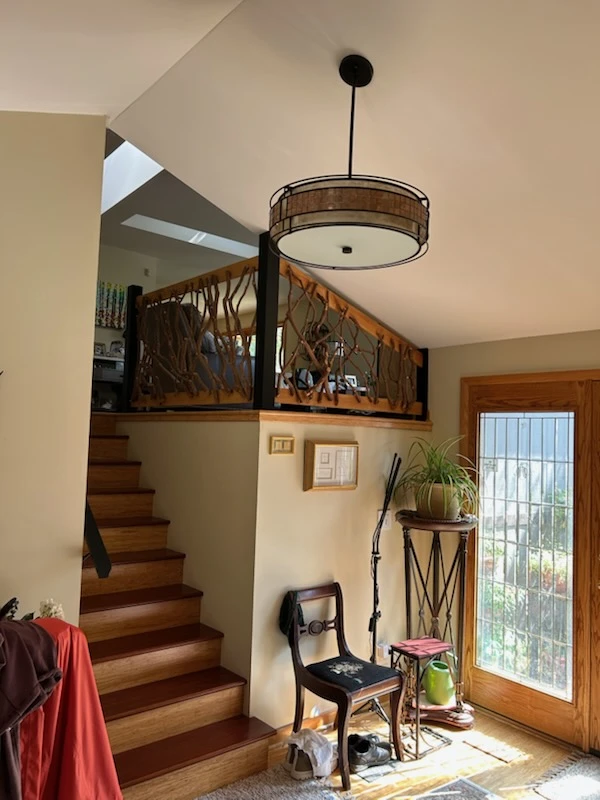 Sunlit entryway with wooden staircase, rustic railing, chair, plants, and ceiling light.
