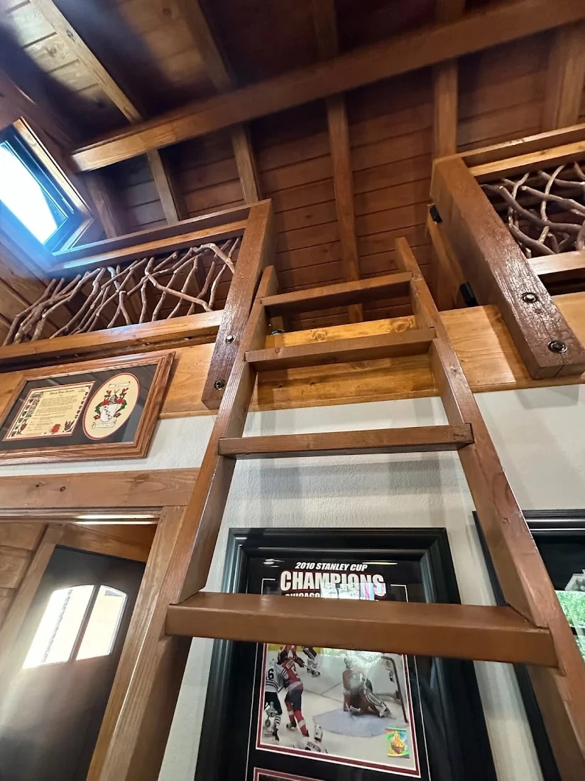 Wooden ladder leading up to a loft with branch-style railing and exposed wood ceiling in a rustic cabin.