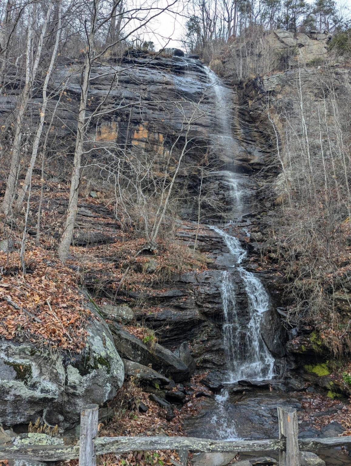 Waterfall cascading down layered rock cliffs in a winter forest with bare trees.